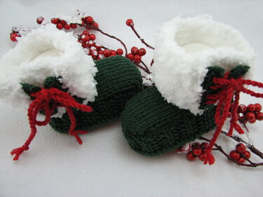 Paar gestrickter dunkelgrüner Babyschuhe mit weißem Umschlag und roten Schnürbändern vor roten Beeren