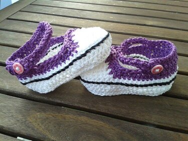 Gehäkelte Babyschuhe in Lila und Weiß im Sneaker-Stil mit rosa Knöpfen auf Holztisch.