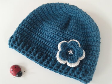 Petrolfarbene gehäkelte Babymütze mit weiß-petrolfarbener Blumenapplikation auf hellem Untergrund