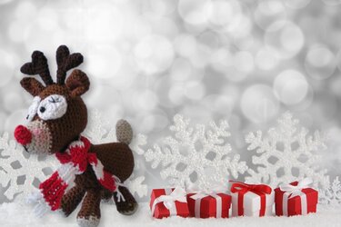 Gehäkeltes Amigurumi-Rentier mit rot-weißem Schal neben roten Geschenkboxen und glitzernden Schneeflocken