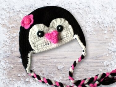 Gehäkelte Pinguin-Mütze in Schwarz-Weiß mit rosa herzförmigem Schnabel, Blume, Ohrenklappen und geflochtenen Bändern