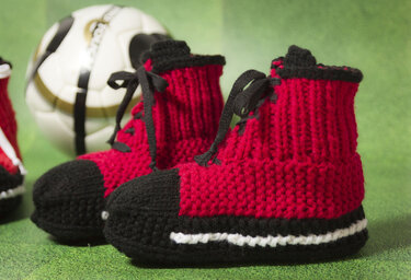 Rote und schwarze gestrickte Babysneaker mit schwarzen Schnürbändern und weißem Streifen vor grünem Untergrund; im Hintergrund ein Fußball.