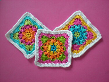Drei bunte gehäkelte Granny Squares mit weißer Umrandung auf rosa Hintergrund