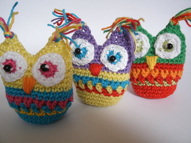 Drei bunte gehäkelte Amigurumi-Eulen mit großen Augen und gestreiften Körpern auf hellem Hintergrund