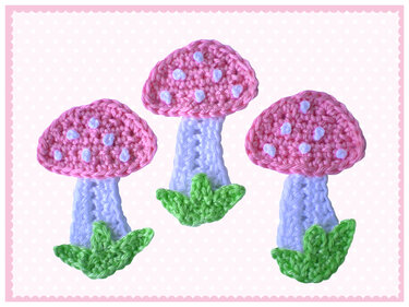 Drei gehäkelte Pilz-Applikationen in Pastellfarben (rosa, hellblau, grün) auf hellem gepunktetem Hintergrund