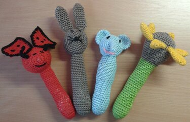 Vier gehäkelte Babyrasseln und Greiflinge auf hellem Holz: rotes Insekt mit schwarzem Rand, grauer Hase, hellblauer Elefant und grau-grüner Greifling mit gelber Blüte.