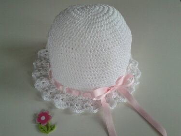 Weiße gehäkelte Babymütze mit gewelltem Rüschenrand und rosa Satinband, daneben kleine Dekoblume