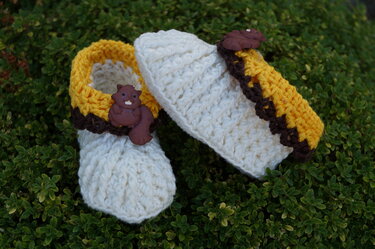 Paar cremefarbener, gelb-brauner gehäkelter Babyschuhe mit braunen Eichhörnchen-Knöpfen auf dunkelgrünem Pflanzenuntergrund