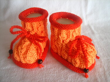 Paar gestrickter Babyschuhe in verschiedenen Orangetönen mit Zopfmuster, roter Kordel und dunklen Perlen