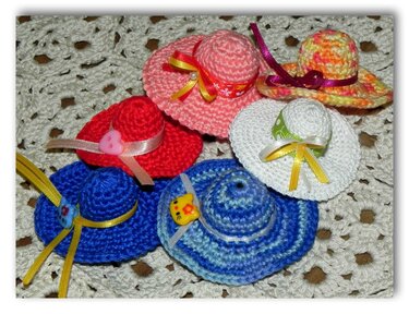 Sechs gehäkelte Mini-Hüte in Pink, Rot, Blau und Weiß, verziert mit Schleifen und kleinen Knöpfen, auf einer hellen gehäkelten Unterlage.