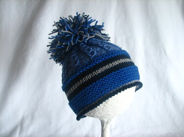Gestrickte Kindermütze mit Bommel in Blau- und Grautönen auf weißem Ständer