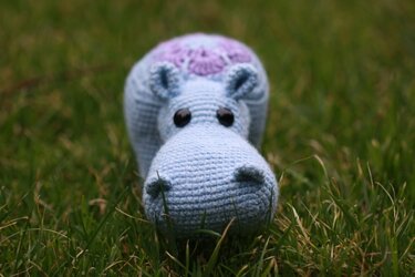 Hellblaues Amigurumi-Nilpferd mit lila Muster und schwarzen Augen auf Gras.