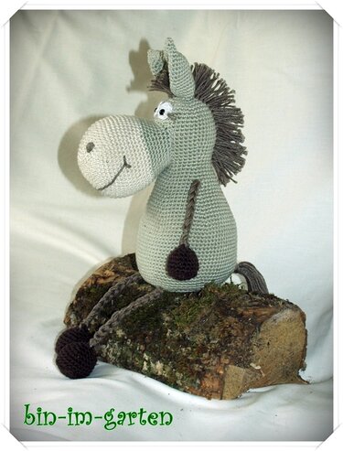 Pastellgrüner gehäkelter Amigurumi-Esel mit brauner Fransenmähne sitzend auf einem Holzstück