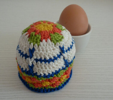 Nahaufnahme eines gehäkelten, mehrfarbigen Eierwärmers neben einem Ei im Eierbecher
