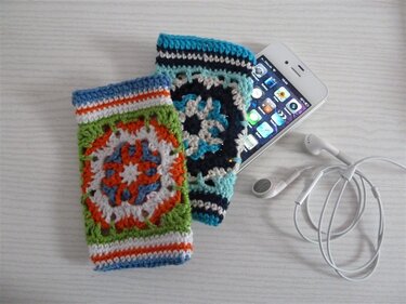 Zwei gehäkelte Smartphone-Hüllen mit rundem Granny-Motiv in bunten Farben, daneben weiße Ohrhörer
