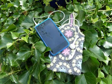 Smartphone mit weißen Kopfhörern auf einer gehäkelten Handytasche im Granny-Square-Muster, umgeben von Efeu.