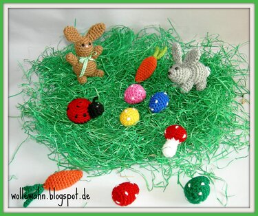 Zwei gehäkelte Hasen, bunte Eier, Karotten, Marienkäfer und Pilze auf grünem Dekogras