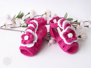 Pink-weiße gehäkelte Babysandalen mit Blüten, arrangiert neben rosa Blütenzweigen.