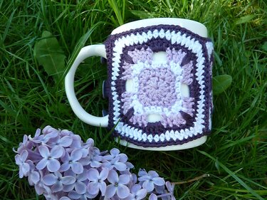 Weiße Tasse mit gehäkelter Granny-Square Hülle in Flieder- und Weißtönen auf Gras mit lila Blüten