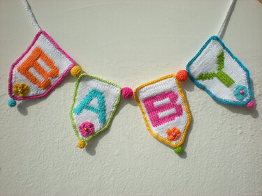 Gestrickte Girlande mit vier weißen Wimpeln, die die Buchstaben BABY in bunten Farben zeigen, verziert mit kleinen Blumen und Kugeln.