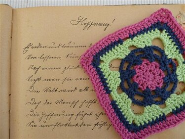 Farbiges Granny Square in Rosa, Grün und Dunkelblau auf den Seiten eines alten Buches.