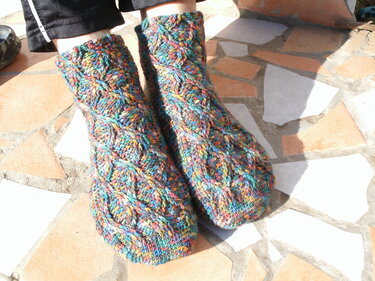 Mehrfarbige, gehäkelte Socken mit Zopfmuster an Füßen auf gefliestem Boden.