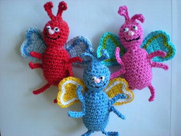 Drei gehäkelte Amigurumi‑Schmetterlinge in Rot, Rosa und Blau mit bunten Flügeln und lächelnden Gesichtern.