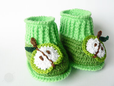 Zwei grüne, gehäkelte Babyschuhe mit applizierten Apfelhälften auf weißem Hintergrund.