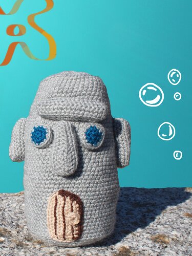Gehäkelte graue Amigurumi-Figur mit blauen Augen und kleiner brauner Tür auf steinigem Untergrund vor türkisem Hintergrund.