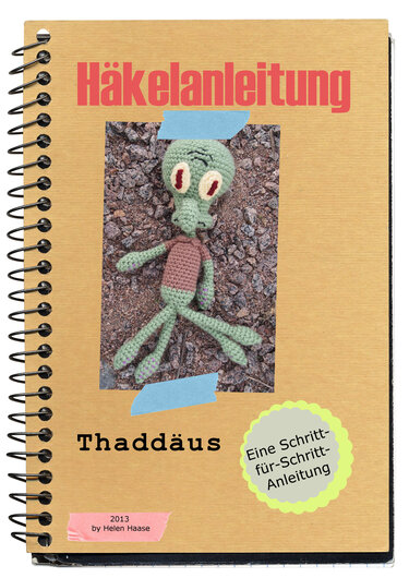 Beigefarbenes Spiralnotizbuch mit dem Schriftzug Häkelanleitung und einer grünen, gehäkelten Krakenfigur auf kiesigem Untergrund.