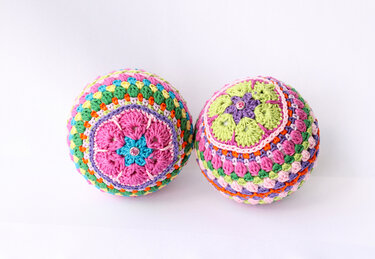 Nahaufnahme von zwei bunten, gehäkelten Kugeln mit Blumenmotiven und kleinen Schmucksteinen.