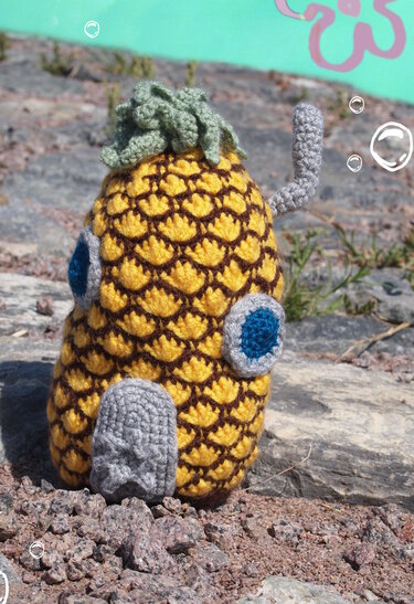 Gehäkeltes Ananas‑Häuschen Amigurumi mit grünem Blatt, runden Fenstern und seitlichem grauen Rohr