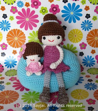 Two crocheted amigurumi dolls, a larger one in purple and a smaller one in pink, seated on a blue pouf with a floral background.