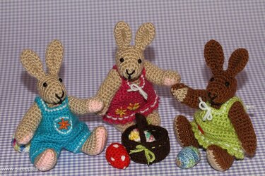 Drei gehäkelte Amigurumi-Hasen in Blau, Pink und Grün mit kleinem Korb und Ostereiern auf lila-kariertem Hintergrund.