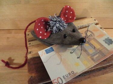 Handgefertigte graue Filzmaus mit rot-weiß gepunkteten Ohren, Perlenaugen und rotem Garnschwanz auf Holzbrett neben Geldschein.