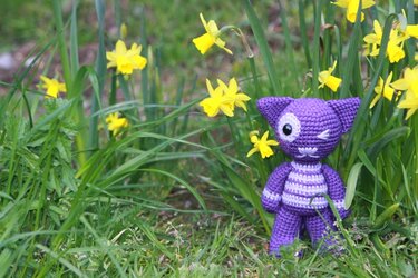 Gestreifte lila Amigurumi-Katze mit Blinzelauge im Frühlingsgras neben gelben Narzissen