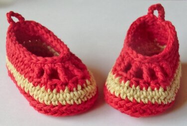 Zwei gehäkelte Babyschuhe in Rot und Gelb auf hellem Hintergrund, seitlich angeordnet