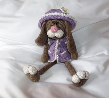 Gehäkelter brauner Amigurumi-Hase mit lila Hut und Kleid auf weißer Oberfläche.