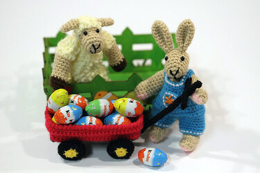 Roter Häkel-Bollerwagen mit bunten Eiern, gezogen von einem Amigurumi-Hasen; dahinter ein Amigurumi-Schaf vor grünem Zaun.