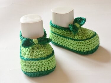 Zwei gehäkelte Babyschuhe in Hellgrün und Dunkelgrün mit Schleifen, auf weißen Fußformen präsentiert.