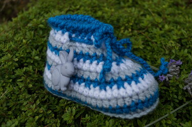 Gehäkelter Babyschuh in Blau, Grau und Weiß mit grauer Hasen-Applikation auf grünem Laub
