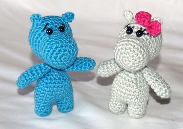 Zwei gehäkelte Amigurumi-Nilpferde, eines blau und eines hellgrau mit rosa Schleife