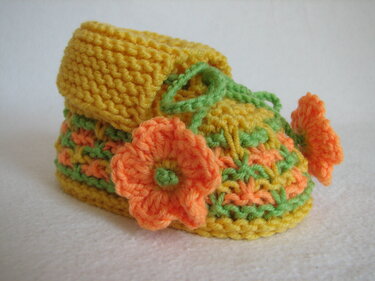 Gestrickter Babyschuh in Gelb, Orange und Grün mit orangefarbener Blume und grünem Band auf hellem Hintergrund.