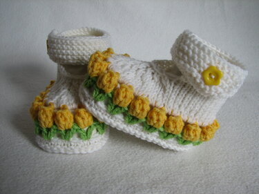 Gestrickte weiße Babyschuhe mit Riemen, gelbem Blütenknopf und gelb-grüner Blumenborte