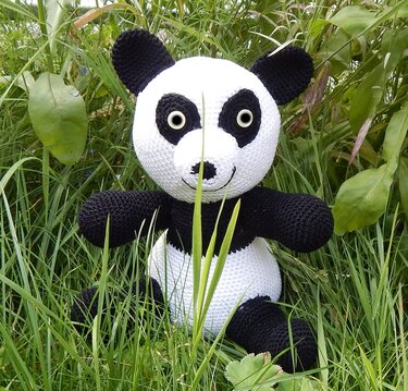 Schwarz-weißer gehäkelter Amigurumi-Panda sitzt im hohen grünen Gras.