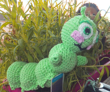 Gehäkelte grüne Raupe Amigurumi mit segmentiertem Körper und großen aufgesetzten Augen vor Pflanzen