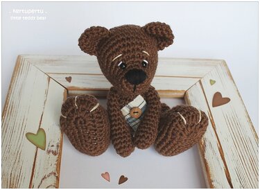 Brauner gehäkelter Amigurumi-Teddybär mit kariertem Patch und rundem Holzknopf in weiß lasiertem Holzrahmen mit Herzmotiven