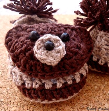 Nahaufnahme gehäkelter Babyschuhe in Braun und Beige mit Bärengesichtern und Pompons auf Korkuntergrund.