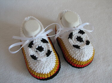 Nahaufnahme zweier weißer, gehäkelter Babyschuhe mit schwarz‑rot‑goldenem Rand und kleinen Fußball‑Applikationen, verziert mit weißen Schleifenbändern.