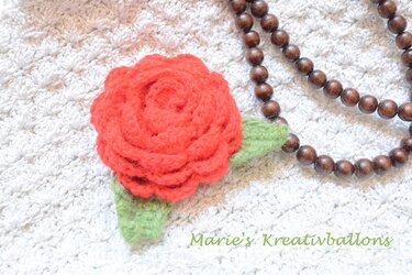 Rote gehäkelte Rose mit grünen Blättern auf weißem Häkelstoff, daneben eine braune Holzperlenkette.